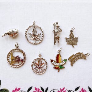May include: A collection of seven silver-toned metal charms featuring Canadian themes. Included are charms shaped like maple leaves, a figure of a Royal Canadian Mounted Police officer, and pieces inscribed with PEI, Thunder Bay, and Canada.