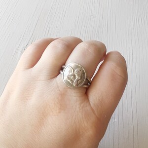 May include: A silver ring with a round design featuring a cherub with wings. The ring is on a person's finger.