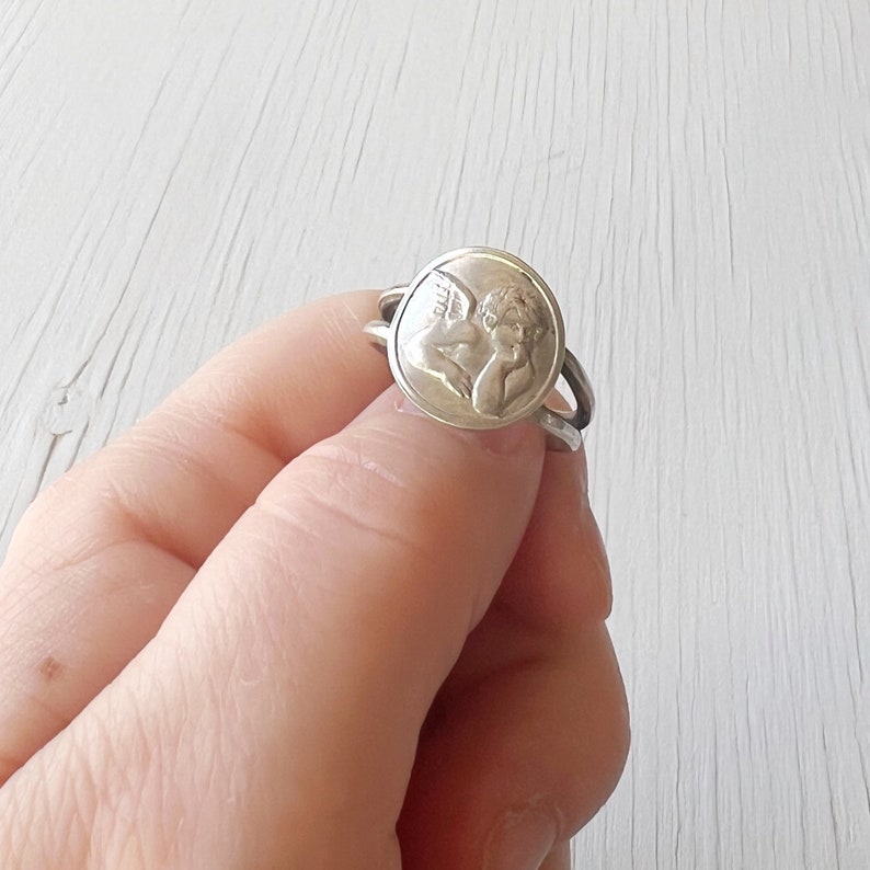 May include: Silver ring with a round, raised design of an angel with wings. The ring is worn on a finger.