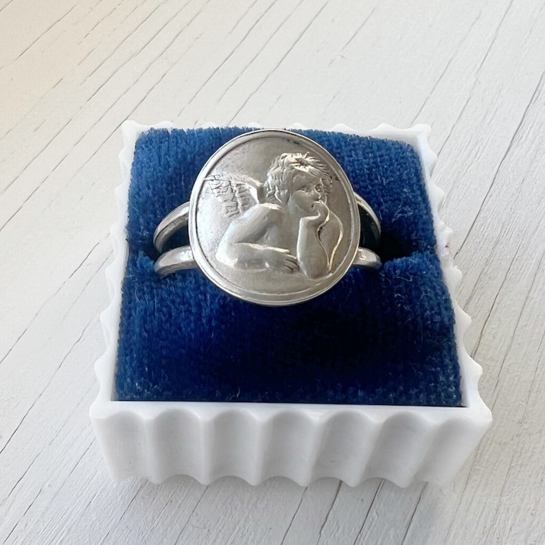 May include: A silver ring with a round design featuring a cherub with wings. The ring is in a blue velvet box.