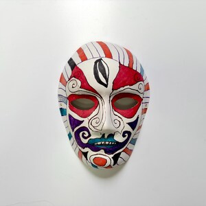 May include: A white face mask featuring hand-drawn designs in red, purple, blue, and black ink. The mask displays symmetrical patterns around the eyes, a central black oval on the forehead, and colorful striped borders along the outer edges.