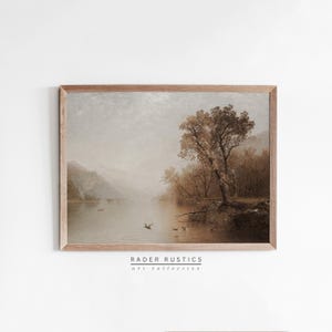 May include: A framed landscape art print featuring a serene lake scene with mountains in the background. The artwork is in muted tones of brown and beige, with a tree on the right side. The print is from Rader Rustics art collective.