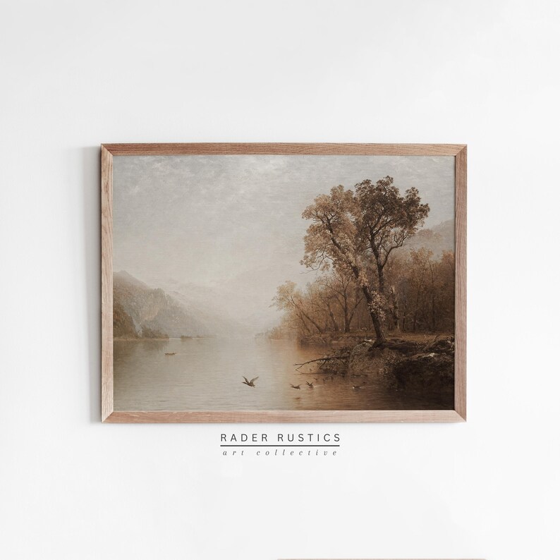 May include: A framed landscape art print featuring a serene lake scene with mountains in the background. The artwork is in muted tones of brown and beige, with a tree on the right side. The print is from Rader Rustics art collective.