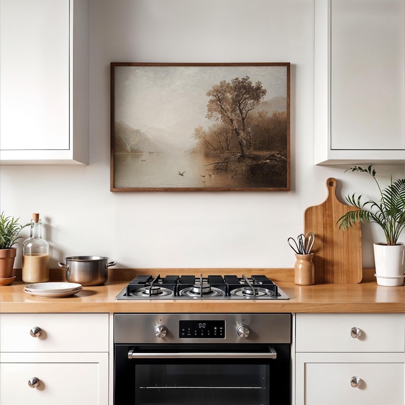 May include: A framed landscape painting depicting a serene lake scene with a large tree on the bank. The artwork features muted tones of brown, beige, and gray, creating a vintage aesthetic. The painting is displayed above a stainless steel oven and stovetop in a kitchen setting.