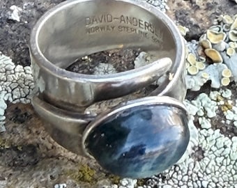 May include: A silver ring with a large, dark blue gemstone. The ring has a textured band and a curved design that cradles the stone. The inscription "DAVID-ANDERSEN NORWAY STERLING 925" is visible on the ring's band.