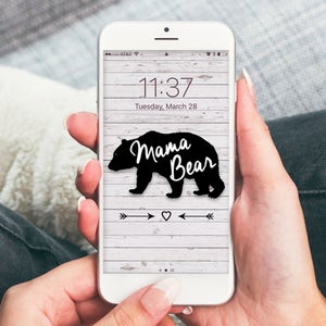 Puede incluir: Un tel&eacute;fono inteligente blanco con una silueta de oso negro y el texto "Mama Bear" en la pantalla. El fondo es un patr&oacute;n de veta de madera con flechas y un coraz&oacute;n.