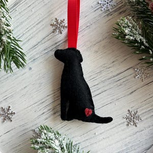 Puede incluir: Adorno de perro de fieltro negro con un corazón rojo en un lado, colgado de una cinta roja.