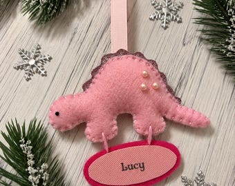 May include: A pink felt dinosaur ornament with a dark pink scalloped spine, three small pearl-like beads, and a light pink ribbon loop for hanging. Attached below is a pink oval tag featuring the name Lucy embroidered in dark gray thread.
