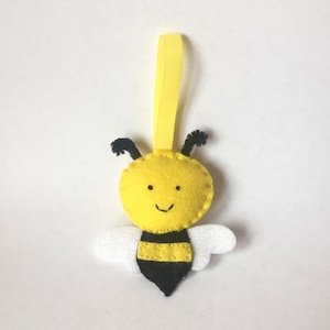 May include: A yellow felt bee ornament with black eyes, a smiling mouth, and black and yellow stripes. The bee has white felt wings and a yellow ribbon for hanging.