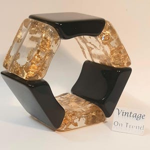 May include: A chunky bracelet with a geometric design. The bracelet features alternating black and clear segments. The clear segments are filled with gold leaf. The bracelet is a statement piece. The text "Vintage On Trend" is in the image.