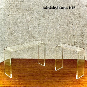 Puede incluir: Dos expositores de acrílico transparente, uno más grande que el otro, sobre una superficie de madera. Los expositores tienen forma de rectángulo con la parte superior e inferior curvadas. Los expositores se utilizan para exhibir artículos como joyas, figuritas u otros objetos pequeños.