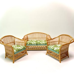 May include: A three-piece wicker furniture set. The set includes a loveseat and two armchairs. The cushions feature a green and white tropical leaf pattern. The wicker is a light brown colour.