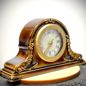 May include: A miniature brown mantel clock with a gold-colored clock face and Roman numerals. The clock has decorative gold accents and a wooden base. The clock is approximately 3 inches wide.