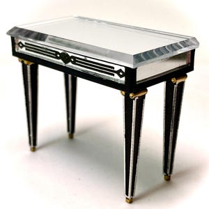 May include: A small black and white table with a clear glass top. The table has gold accents on the legs and a geometric design on the front.