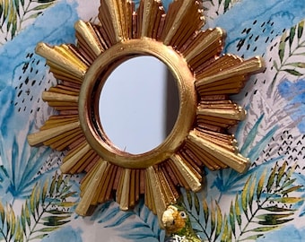 May include: A gold sunburst mirror with a round, reflective surface. The mirror is set against a blue and green tropical print background.