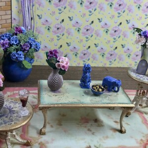 以下が含まれることがあります： 花をテーマにしたミニチュアのシーン。青いセラミックの象の置物、金のビーズのボウル、紫の花の青いセラミックの花瓶が置かれた小さなテーブル。紫の花の青いセラミックの花瓶と花柄の青いセラミックの花瓶が置かれたもう1つのテーブル。テーブルは模様のある敷物の上に置かれています。