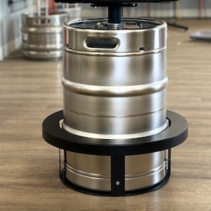 May include: A stainless steel keg with a black metal base and a ring around the top. The keg is sitting on a wooden floor.