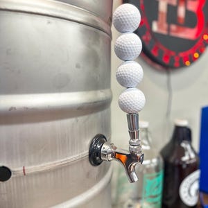 Puede incluir: Un barril de cerveza de acero inoxidable con un grifo con un mango de pelota de golf. El mango del grifo está hecho de cuatro pelotas de golf blancas apiladas una encima de la otra. El barril tiene la palabra "TAB".