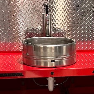 May include: A custom bar sink made from a stainless steel keg. The sink is set into a red cabinet with drawers, and a chrome tap is mounted above. The background features a diamond-plate metal backsplash.