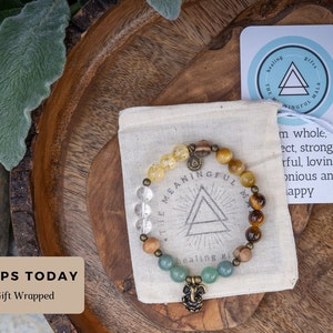 May include: A beaded bracelet with a variety of stones, including clear quartz, tiger's eye, and green aventurine. The bracelet has a metal charm in the shape of a triangle with the words "The Meaningful Mala" inscribed on it. The bracelet is packaged in a small white pouch.