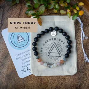 May include: Black beaded bracelet with a silver charm that says "The Light Within". The bracelet is in a small white drawstring bag with a black and white logo that says "The Meaningful Mala".