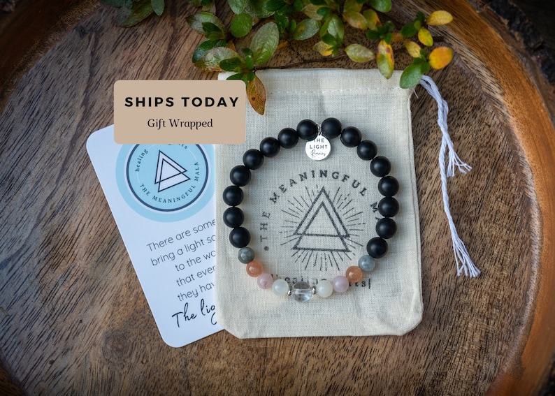 May include: Black beaded bracelet with a silver charm that says "The Light Within". The bracelet is in a small white drawstring bag with a black and white logo that says "The Meaningful Mala".