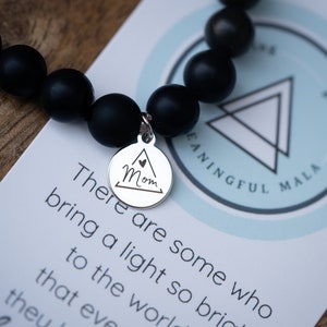 May include: Black beaded bracelet with a silver charm that says "Mom" inside a triangle. The bracelet is on a white card with the text "There are some who bring a light so bright to the world that even theu" and a logo that says "MEANINGFUL MALA".