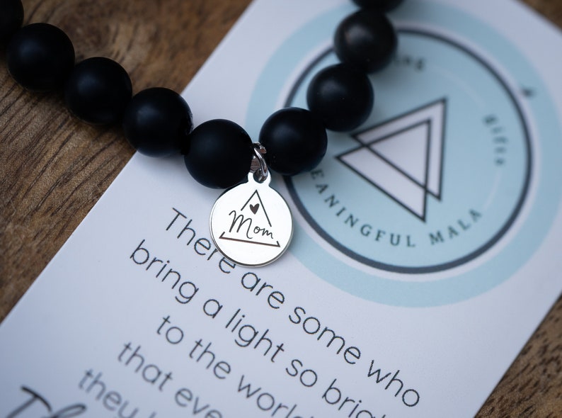 May include: Black beaded bracelet with a silver charm that says "Mom" inside a triangle. The bracelet is on a white card with the text "There are some who bring a light so bright to the world that even theu" and a logo that says "MEANINGFUL MALA".