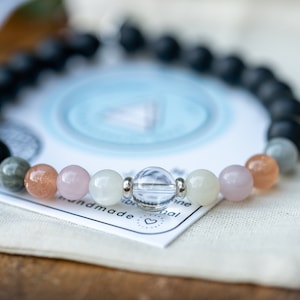 May include: A bracelet made with black, grey, white, pink, and orange beads. The bracelet is on a white fabric surface with a blue and white card that says "handmade" and "original".