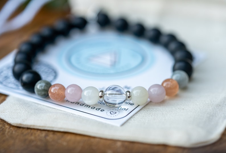 May include: A bracelet made with black, grey, white, pink, and orange beads. The bracelet is on a white fabric surface with a blue and white card that says "handmade" and "original".