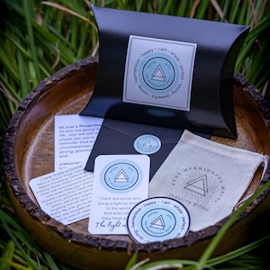 May include: A set of three small, round stickers with a blue and white design. The stickers feature a triangle with the text "The Meaningful Mala" inside. The text "Healing Gifts" is printed on the sticker. The stickers are on a wooden surface with green grass in the background.