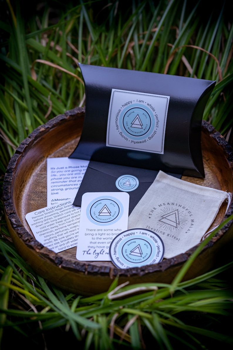 May include: A set of three small, round stickers with a blue and white design. The stickers feature a triangle with the text "The Meaningful Mala" inside. The text "Healing Gifts" is printed on the sticker. The stickers are on a wooden surface with green grass in the background.