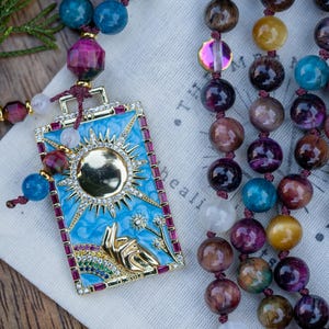 May include: A gold pendant featuring a blue and white enamel design of a sun and a hand reaching upwards. The pendant is attached to a long necklace made of various coloured gemstones, including tiger's eye, amethyst, and clear quartz.