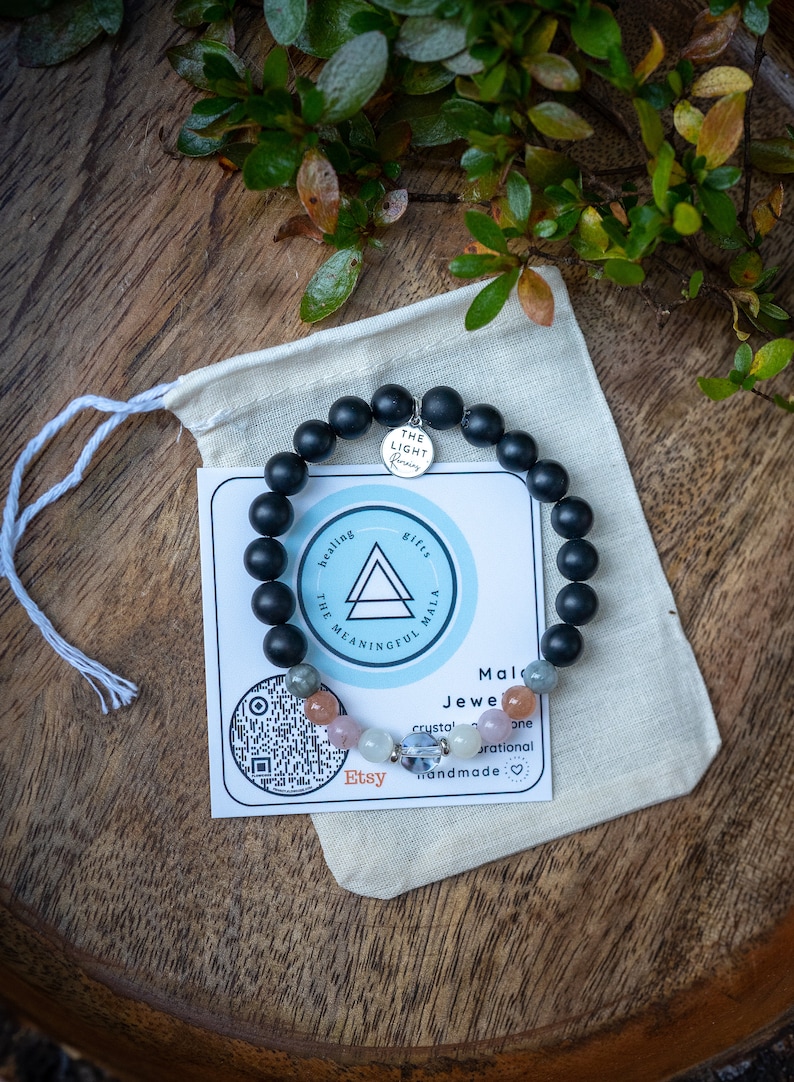 May include: Black beaded bracelet with a silver charm that says "The Light Within". The bracelet is on a white fabric pouch with a card that says "Healing, The Meaningful Mala, Gifts" and "Mala Jewelry, Crystal, One Rational, Handmade".