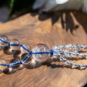 May include: A clear quartz crystal bead bracelet with blue accents. The beads are strung on a blue cord, and the bracelet is displayed on a wooden surface. The bracelet features a larger central bead.
