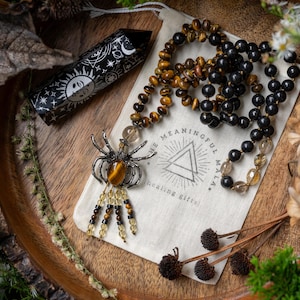 Puede incluir: Un collar de cuentas negras y ojo de tigre con un dije de araña de plata y un dije de cristal. El collar se muestra en una bolsa de tela blanca con el texto "The Meaningful Mala" y "healing gifts".