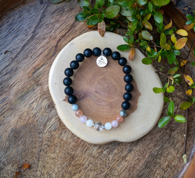 May include: A black beaded bracelet with a silver charm that says "Mom" and a mix of light pink, gray, and white beads.