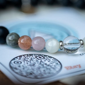 May include: A bracelet made of various stones, including black, grey, pink, white, and clear. The bracelet is on a white card with a black and white design and the word "Play" in red.