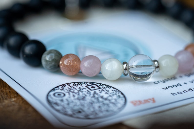 May include: A bracelet made of various stones, including black, grey, pink, white, and clear. The bracelet is on a white card with a black and white design and the word "Play" in red.