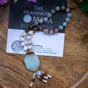 May include: A beaded necklace featuring a large, light blue, raw-cut stone pendant. The necklace is composed of round beads in shades of white, brown, and light blue, arranged in a circular pattern. A small tassel of beads hangs from the bottom of the pendant, which rests on a white card with the text THE MEANINGFUL MALA.