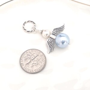 May include: A silver-toned angel charm with a light blue pearl body and detailed wings. The charm includes a silver ring for attachment. A U.S. dime is next to the charm for size comparison.