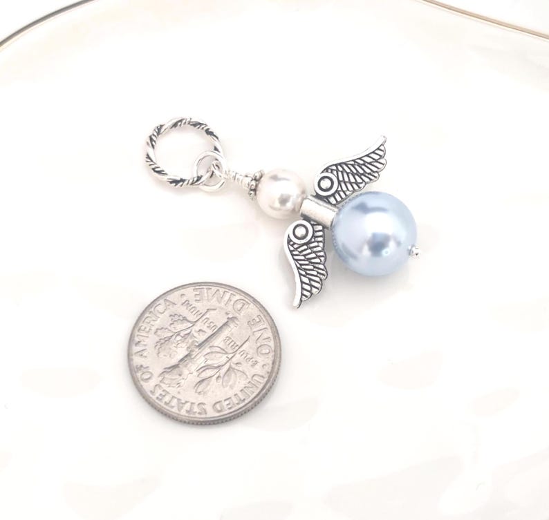 May include: A silver-toned angel charm with a light blue pearl body and detailed wings. The charm includes a silver ring for attachment. A U.S. dime is next to the charm for size comparison.