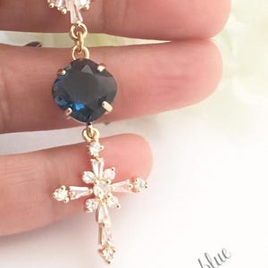May include: A gold-toned pendant featuring a top fan-shaped clear crystal accent, a central cushion-cut dark blue stone, and a lower cross-shaped charm adorned with multiple clear baguette and round crystals, suspended from a delicate gold chain.