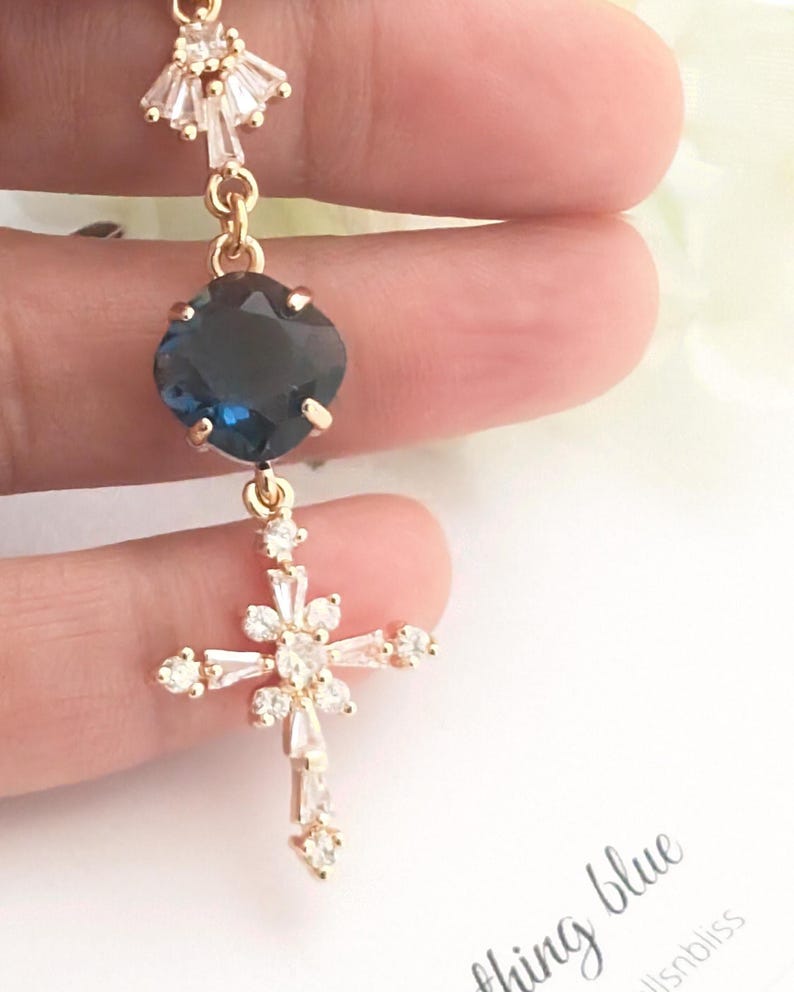 May include: A gold-toned pendant featuring a top fan-shaped clear crystal accent, a central cushion-cut dark blue stone, and a lower cross-shaped charm adorned with multiple clear baguette and round crystals, suspended from a delicate gold chain.