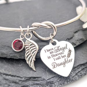 Op de afbeelding: Zilverkleurige armband met een knoopontwerp. Bedels zijn een rode edelsteen, een vleugel en een hartvormige hanger met de tekst "Ik heb een engel in de hemel, ik noem haar dochter."