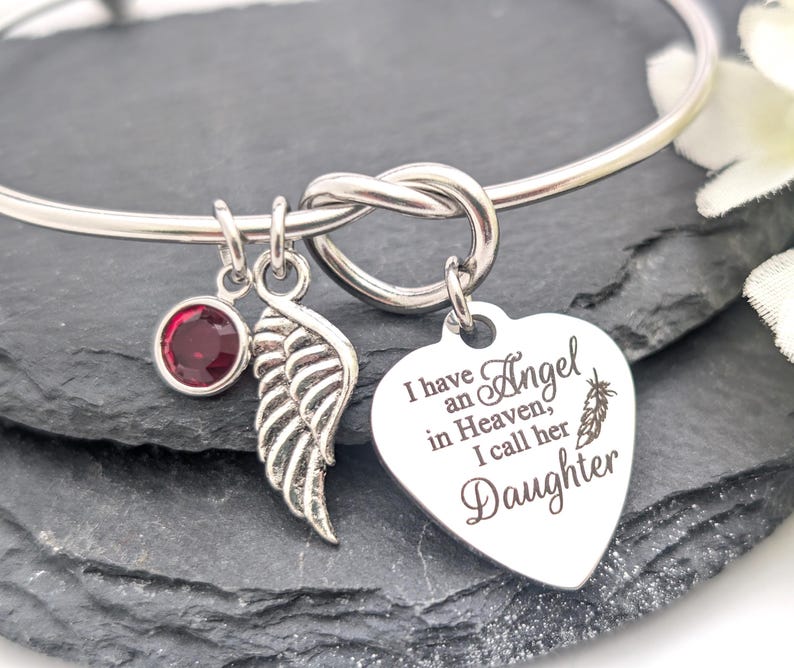 Op de afbeelding: Zilverkleurige armband met een knoopontwerp. Bedels zijn een rode edelsteen, een vleugel en een hartvormige hanger met de tekst "Ik heb een engel in de hemel, ik noem haar dochter."