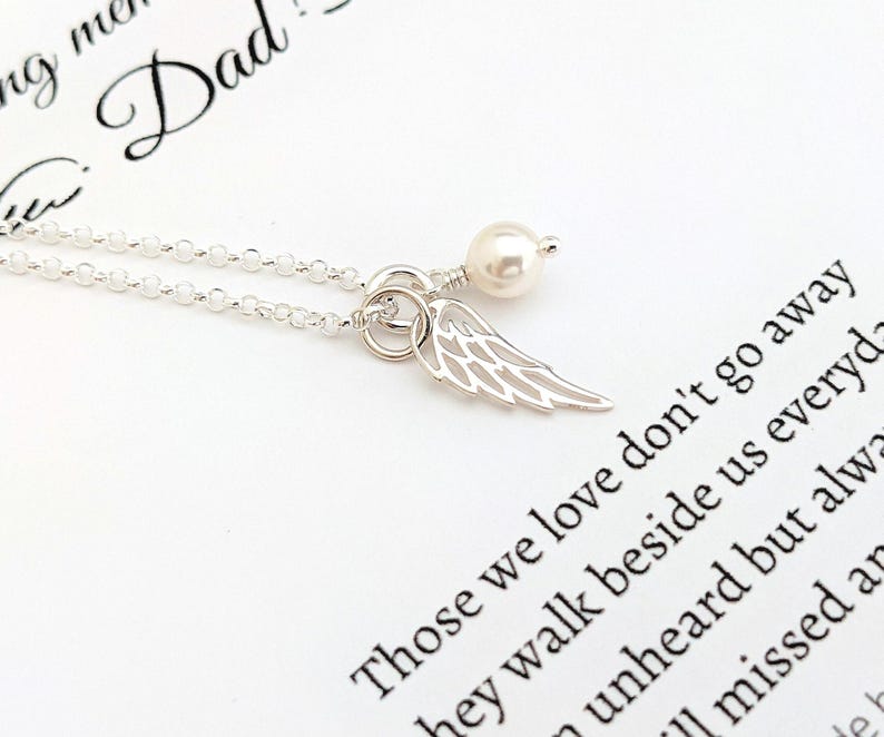 May include: A silver necklace with a delicate chain, featuring a small, openwork wing charm and a pearl. The necklace is displayed on a white surface with printed text that reads "Those we love don't go away".
