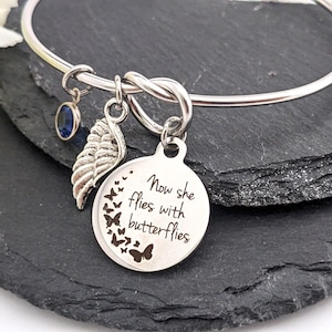 May include: Silver bangle bracelet with a charm that reads "Now she flies with butterflies". The charm has a silver wing and a blue crystal charm.