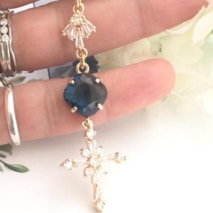 May include: A gold-toned pendant featuring a small fan-shaped crystal accent at the top, a central square-cut dark blue gemstone, and a lower cross-shaped charm encrusted with clear baguette and round-cut crystals, all linked by gold-toned metal.