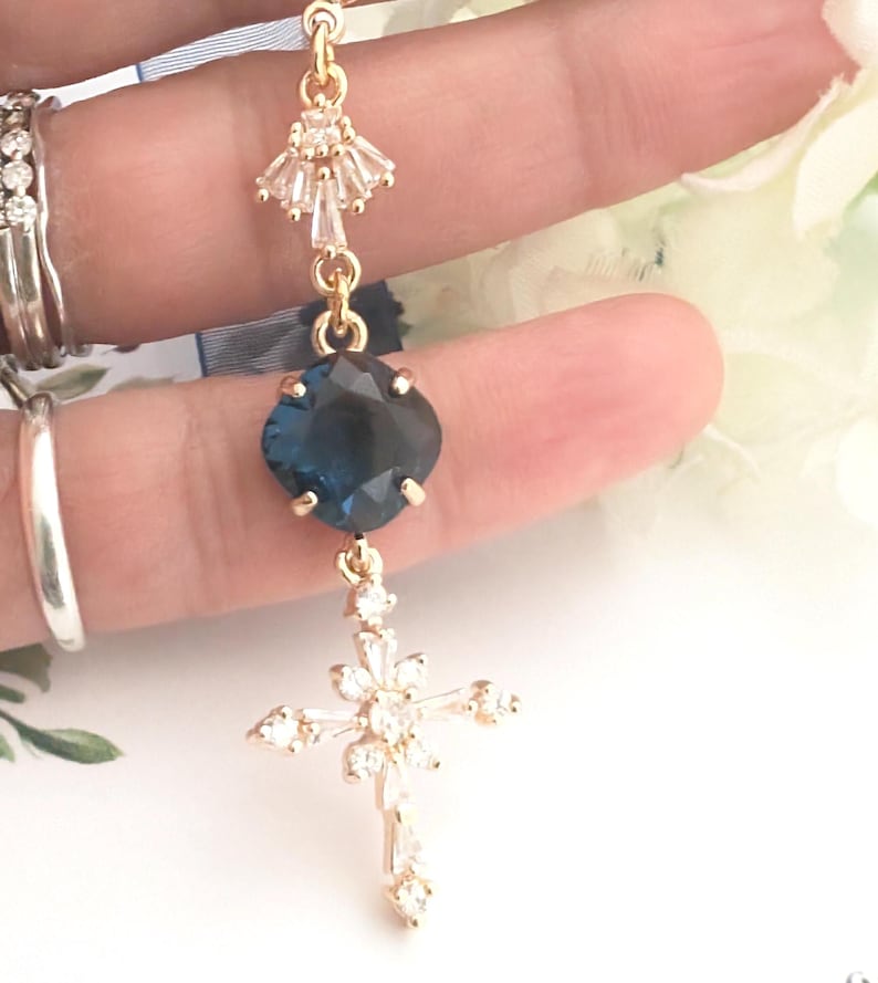 May include: A gold-toned pendant featuring a small fan-shaped crystal accent at the top, a central square-cut dark blue gemstone, and a lower cross-shaped charm encrusted with clear baguette and round-cut crystals, all linked by gold-toned metal.